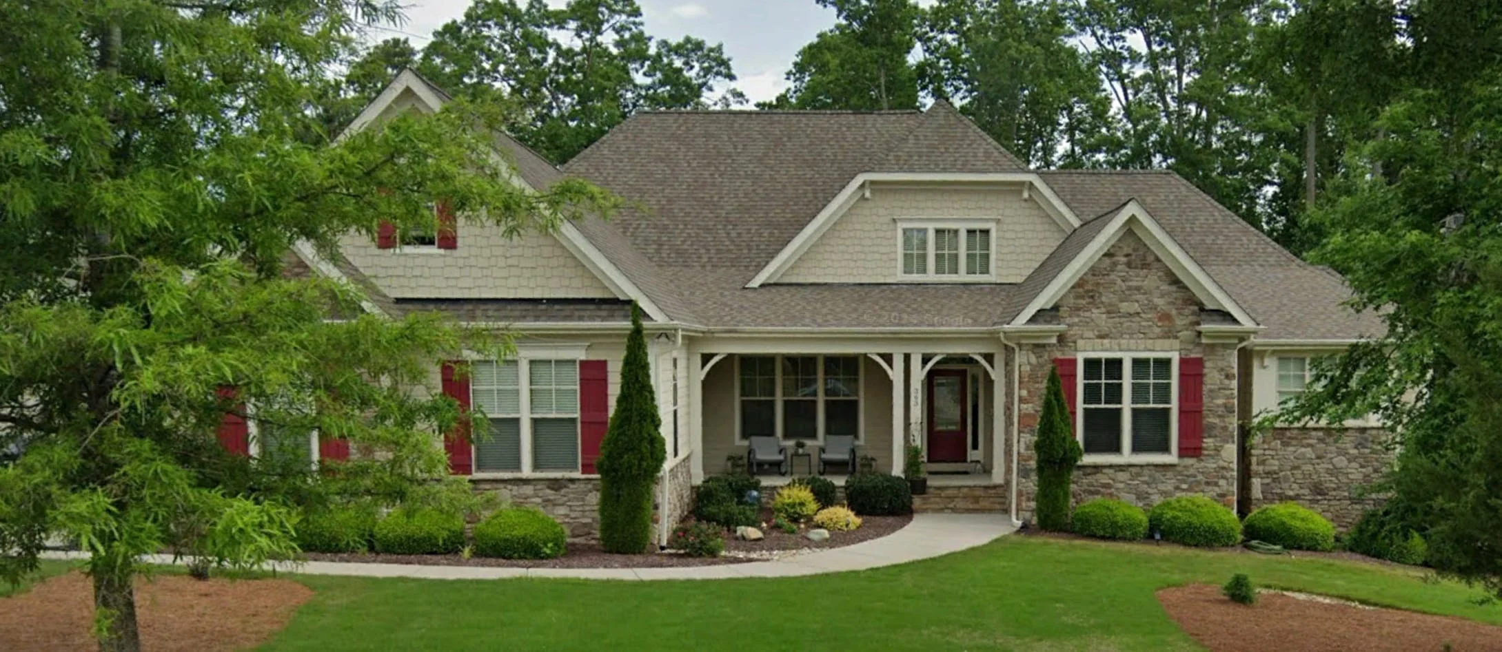 Craftsman cottage-style home with stone accents, red shutters, and front porch seating surrounded by lush landscaping.